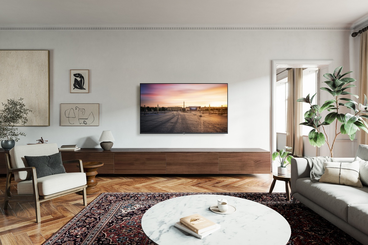 Panasonic W90A na ścianie, na grafice producenta. Telewizor wisi w salonie pełnym mebli i dywanem