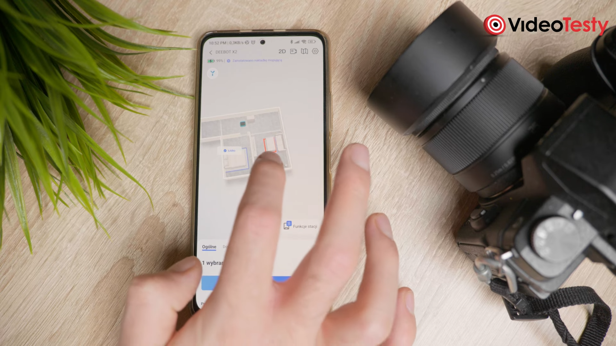 Mężczyzna korzysta z aplikacji Ecovacs Deebot X2 Omni na telefonie znajdującym się na stole, koło sztucznej rośliny i aparatu