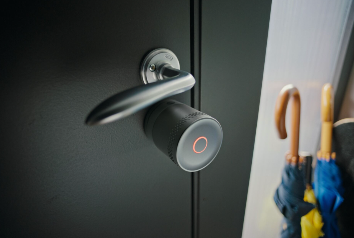 Srebrna wersja zamka Yale Linus Smart Lock L2 Lite na drzwiach. Zamek jest cylindryczny, a drzwi czarne. W tle parasol