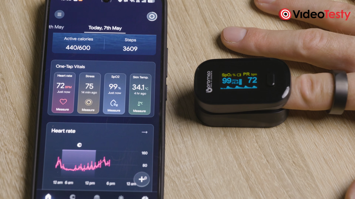 Widok na dloń, na której znajduje się pulsoksymetr oraz ciemny pierścień. Obok jest telefon. Aplikacja w smartfonie oraz pulsoksymetr pokazują te same wyniki