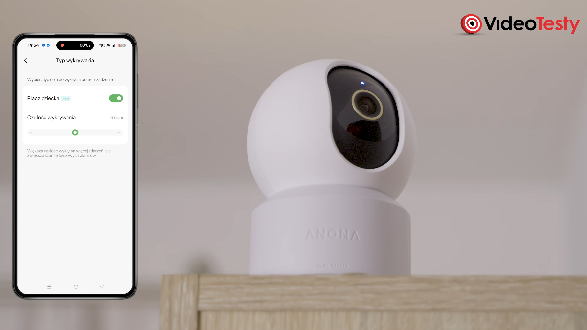 Widok na mockup telefonu z aplikacją Anona, na której ustawia się typ wykrywania płaczu dziecka. Obok widać biało-czarną kamerę z ledwo widocznym napisem Anona na podstawie