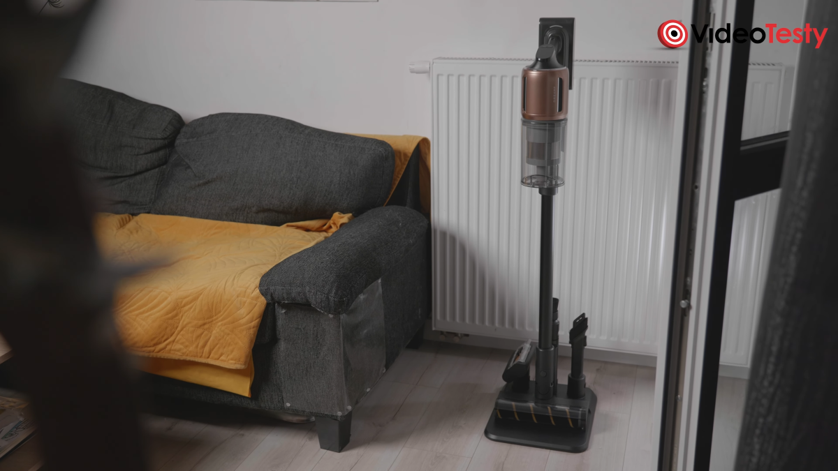 Dreame Z20 widziany w stacji z daleka. Po lewej stronie sofa z żółtą narzutą, po prawej lekko otwarte drzwi balkonowe