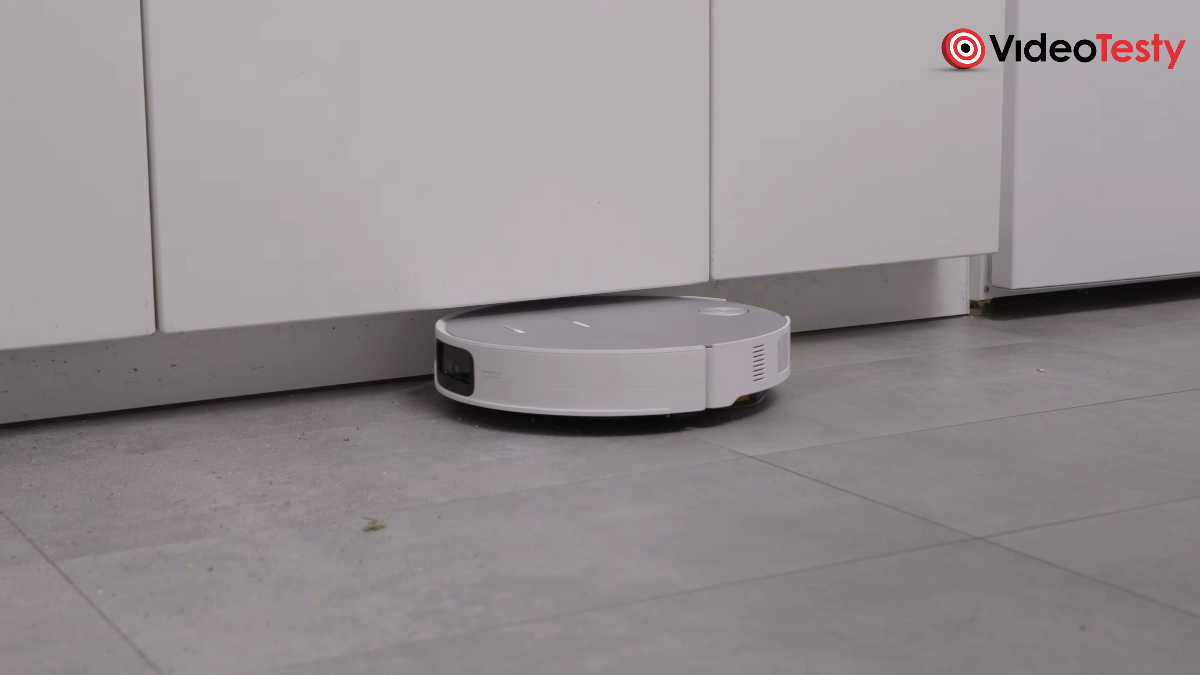Biały okrągły robot przy krawędzi pod nawisem białych mebli kuchennych, czyści kafelki