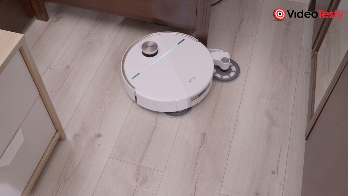 Biały robot wysuwa okrągły dysk mopujący, sprzątając drewnianą podłogę. Dojeżdża do ciemnej, drewnianej szafki