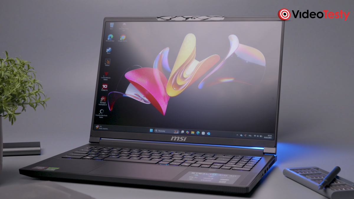 Ciemny laptop widziany pod kątem na szarym tle. Na ekranie ma kolorową tapetę, obok niego znajduje się kilka rzeczy