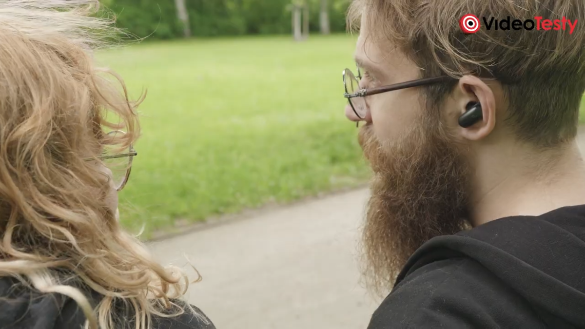 Mężczyzna i kobieta koło siebie. Mężczyzna ma słuchawki oraz okulary, kobieta blond włosy. Siedzą na ławce w parku. Kamera jest za nimi