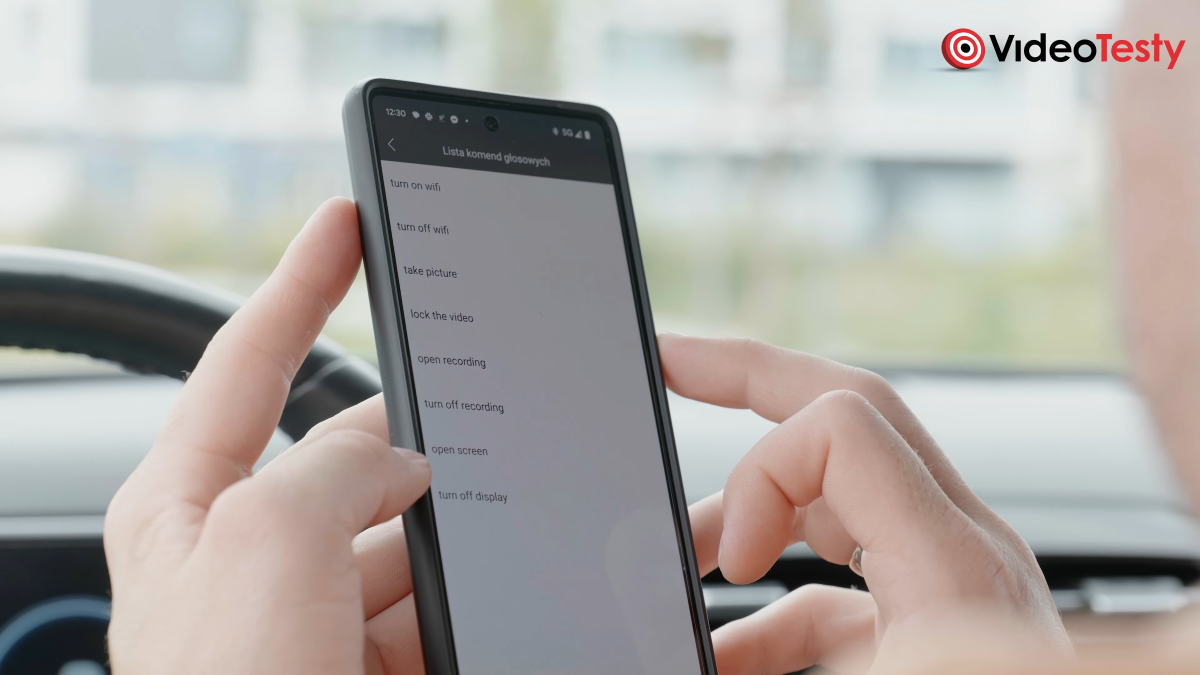 Zbliżenie na komendy głosowe w aplikacji. Wyświetlane są na telefonie w męskich rękach. W tle kierownica