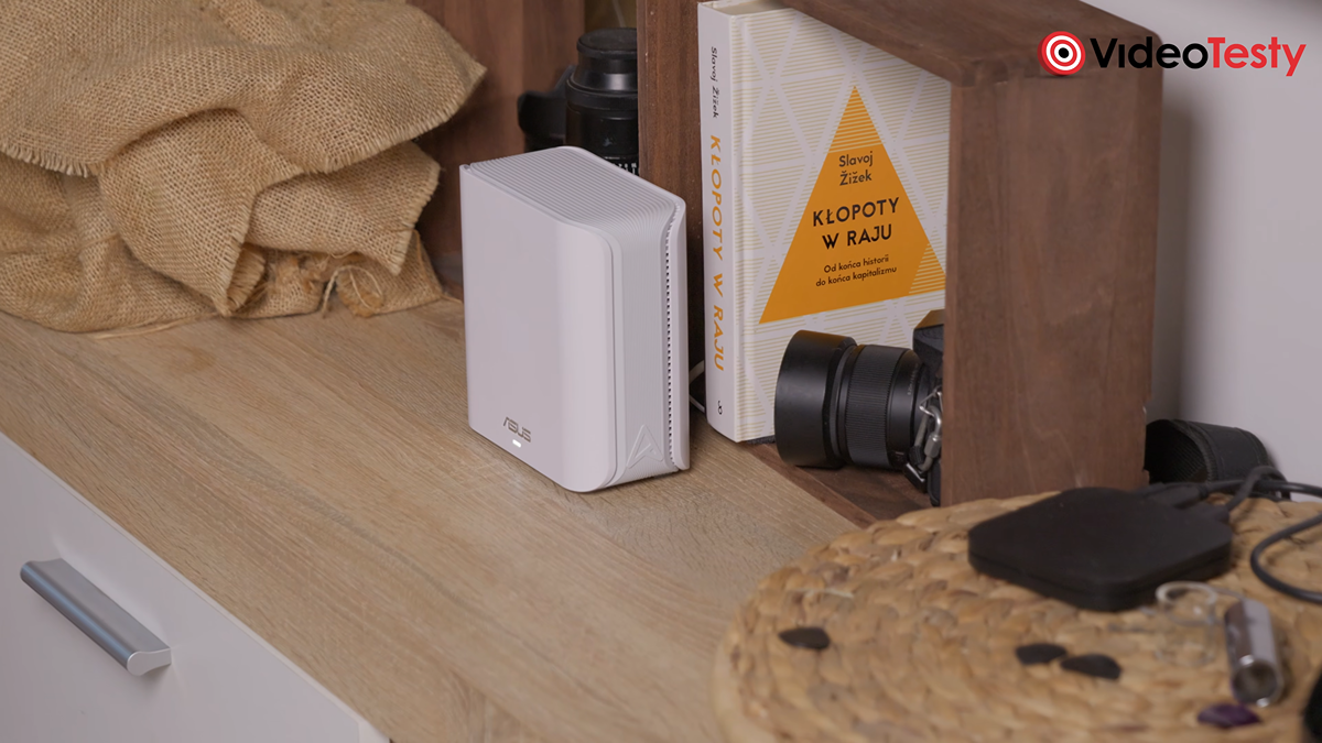 Widok z daleka na biały router stojący na beżowym regale koło książki Kłopoty w raju Slavoya Zizka oraz różnych bibelotów
