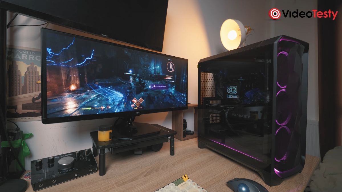 Gra w Dragon Age: widok na biurko z monitorem, na którym znajduje się gra. Obok monitora czarny komputer z fioletowo podświetlonymi wentylatorami i szklanym bokiem