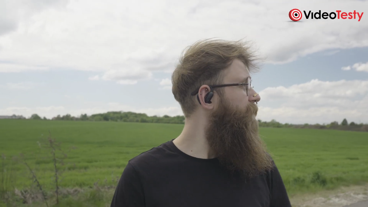 Widok na mężczyznę z czarnymi słuchawkami Soundcore AeroFit 2 Pro na uchu na dworze, z zielonym polem z tyłu oraz chmurami