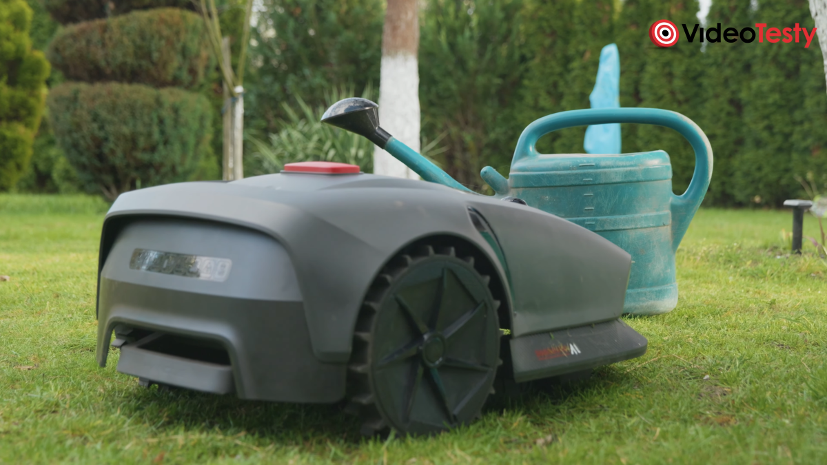 Szaro-czarny robot koszący na tle ogrodu koło zielonej konewki