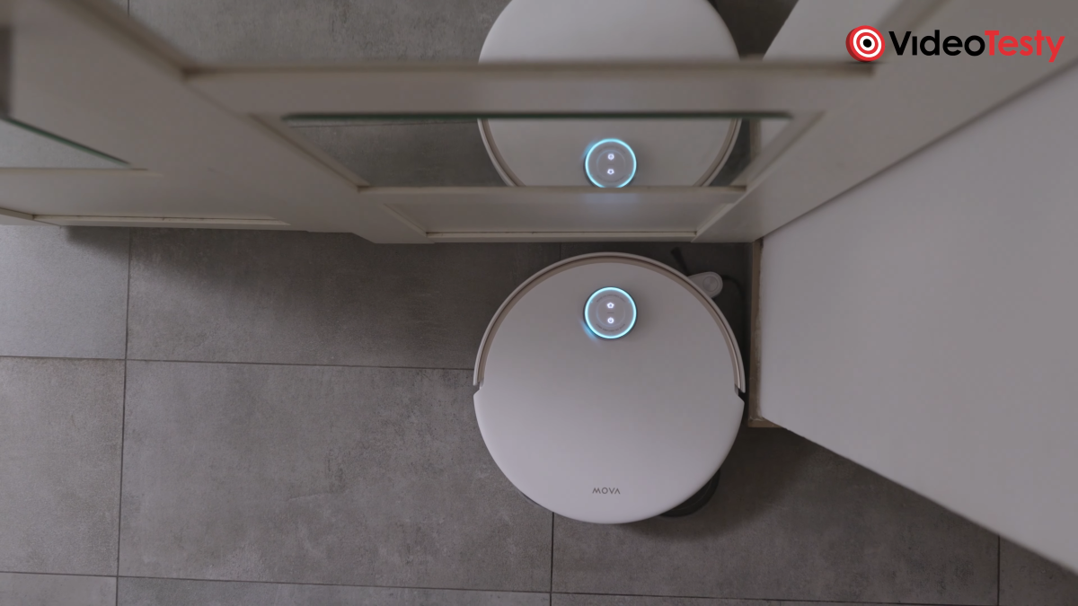 Biały robot z niebiesko podświetlonym LiDAR-em przed szafą z lustrami sprzata róg