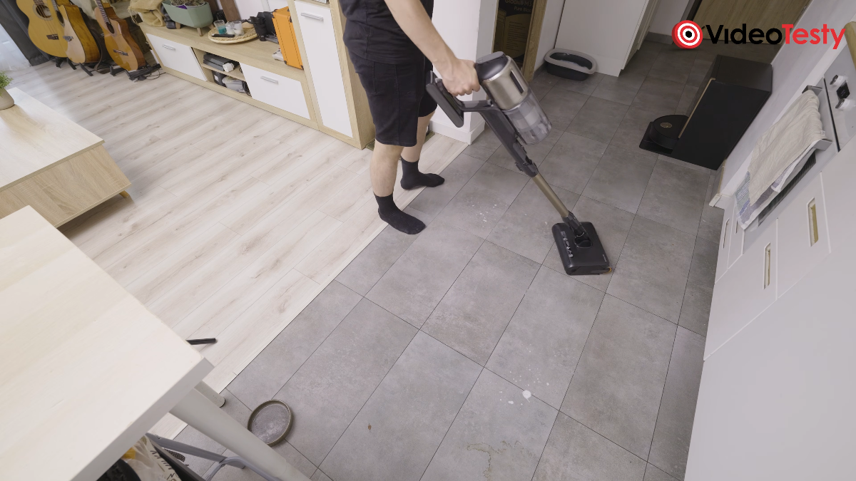 Mężczyzna myje kafelki w pokoju przy pomocy szarego odkurzacza z końcówką do mopowania