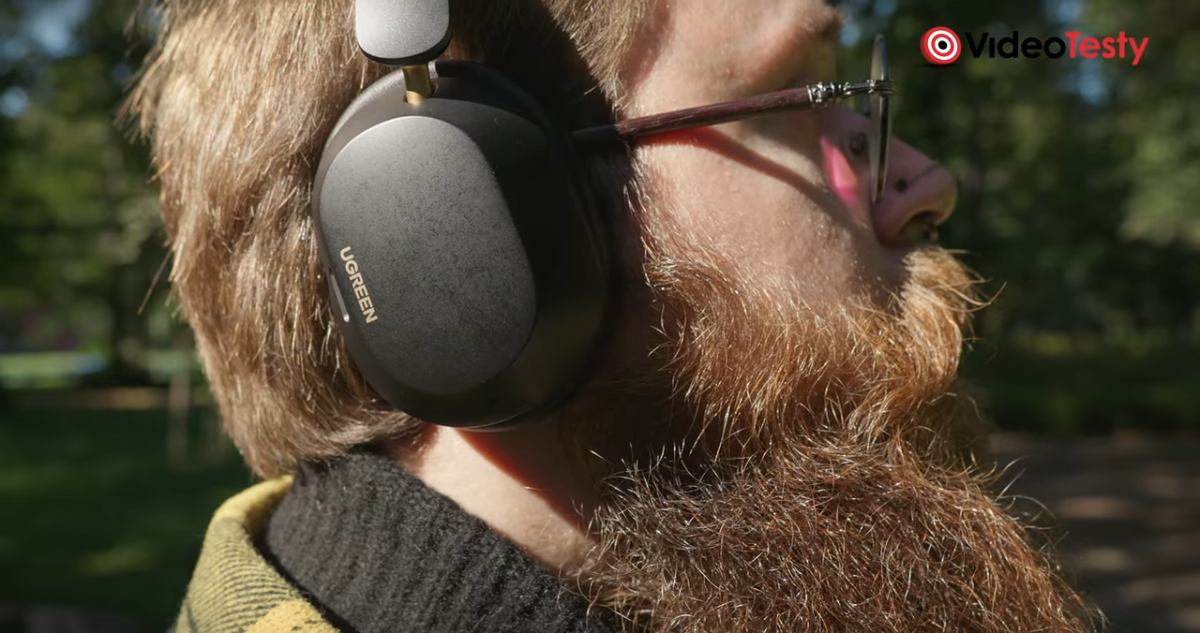 Mężczyzna słuchający muzyki z czarnych słuchawek. Mężczyzna ma okulary i jest brodaty, stoi w parku, zbliżenie na głowę