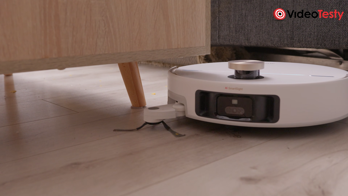 Biały robot wysuwa szczotkę obrotową, doczyszczając podłogę wokół mebla