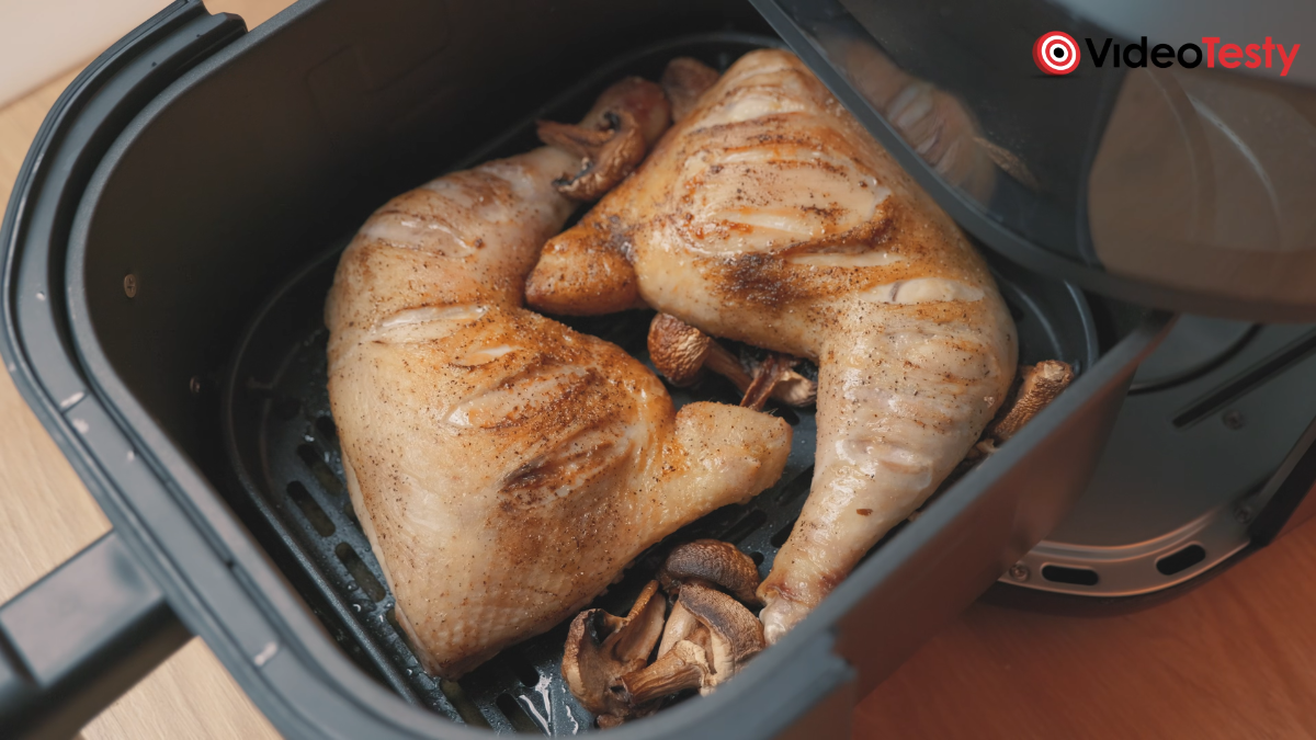 Widziane od góry w czarnym koszu frytkownicy ćwiartki z kurczaka. Są dobrze wypieczone i miejscami nacięte. Wokół nich pieczone pieczarki.